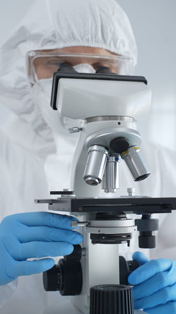 Male virologist in a hazmat suit, mask, and blue gloves, diligently working at a microscope in a...