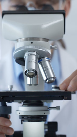 Female scientist wearing lab coat and protective glasses using a modern microscope in a laboratory...