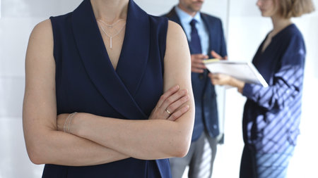 Professional businesswoman stands confidently with arms crossed in sharp focus as colleagues interact behind her during an office meeting, symbolizing leadership and corporate successの写真素材