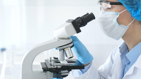 Female scientist analyzing research sample through microscope, wearing protective lab gear for...