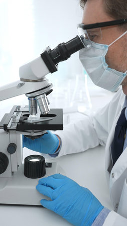 Man scientist wearing protective googles and blue gloves, is using microscope analyzing samples in...