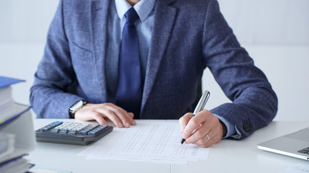 Businessman wearing suit and tie is using calculator and taking notes while working at desk in office, with laptop and stack of folders nearby, close up. Audit and taxes theme in businessの写真素材