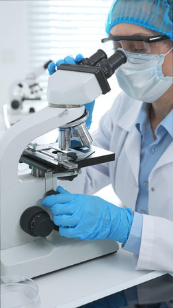 Female scientist analyzing sample through microscope, wearing protective lab gear during medical...