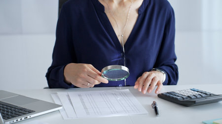 Professional female accountant analyzing financial documents, using magnifying glass and calculator at workplace desk, focused on detailed accounting review, close up. Audit and taxes in businessの写真素材