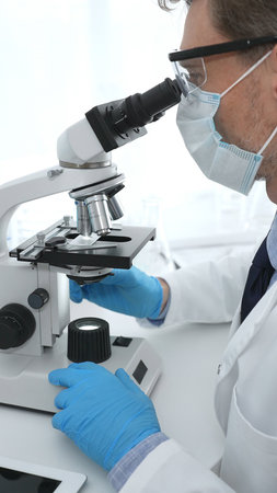 Man scientist wearing protective googles and blue gloves, is using microscope analyzing samples in...