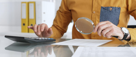 Accountant analyzing financial data with magnifying glass, calculating expenses, and auditing documents at office desk. Audit and taxes in businessの写真素材