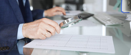 Professional male hand holding magnifying glass, looking at financial data on paper, checking details in accounting and finance. Topic of audit and taxes in businessの写真素材