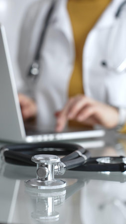 Stethoscope and clipboard with medical documents are on the table near female doctor working on laptop, managing patient data in clinic. Concept of medicine, healthcare and scienceの写真素材