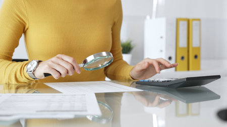 Woman auditing financial documents using magnifying glass and calculator, checking details, managing finance in office. Audit and taxes in businessの写真素材
