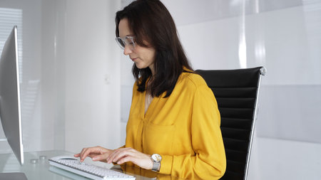 Businesswoman wearing yellow casual blouse and glasses is typing on laptop at office glass desk, analyzing data for business tasks. Audit in businessの写真素材