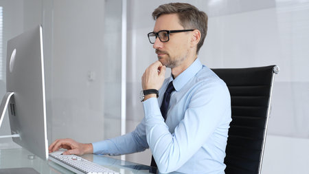 Businessman sitting at a clean office desk, wearing glasses and a light blue shirt with a tie,...