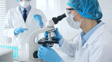 Woman scientist in blue protective gear is examining specimen under microscope while laboratory...