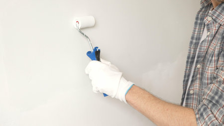 Male painters gloved hand gripping paint roller, spreading white paint across wall during home interior transformation with professional precision, close-up view. Renovation conceptの写真素材