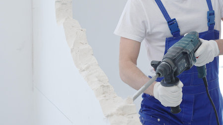 Male construction worker demolishing white wall with rotary hammer drill, wearing blue overalls and protective white gloves, generating dust during renovation project, closeup viewの写真素材