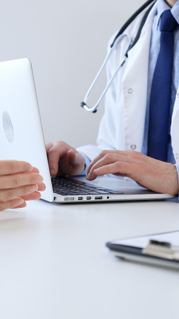 Male doctor is taking notes with laptop while listening to a female patient explaining her symptoms during a medical consultation in the clinic office. Medicine and health careの写真素材