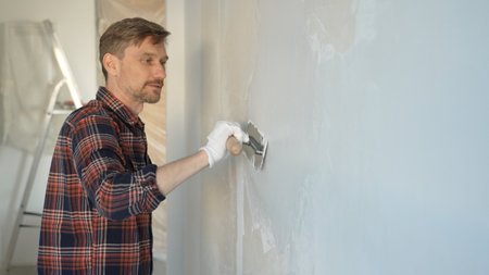Male construction worker wearing gloves is carefully applying plaster to a wall using a hand tool, contributing to a home renovation or construction projectの写真素材