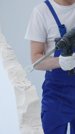 Construction worker demolishing white wall with rotary hammer drill, wearing blue overalls and protective white gloves, generating dust during renovation project, closeup viewの写真素材