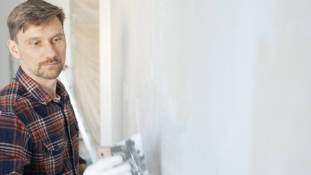 Male construction worker spreading plaster on white wall, wearing protective gloves during home renovation project with putty knifeの写真素材
