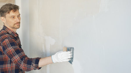 Construction worker man wearing gloves and holding a putty knife is applying plaster on a white wall, performing home renovation and improvementの写真素材