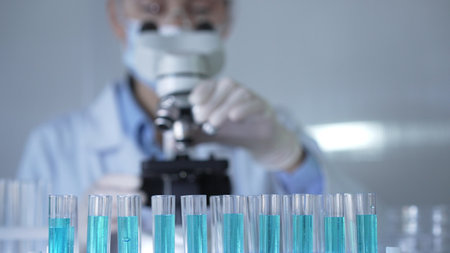 Laboratory tubes, filled with a blue liquid, are in focus, closeup view. Female scientist researcher wearing white gloves and protective mask is using a microscope at the backgroundの写真素材