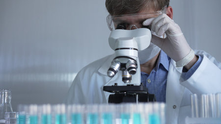 Male scientist, wearing a lab coat, mask, white gloves and safety glasses, is using a binocular...