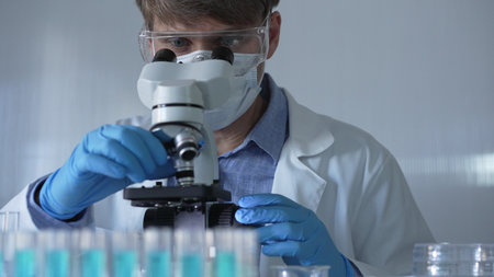 Scientist man, wearing a lab coat, mask, blue gloves and safety glasses, is using a binocular...