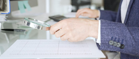 Male accountant holding magnifying glass, checking financial documents on office desk, analyzing taxes and data. Audit and taxes in businessの写真素材