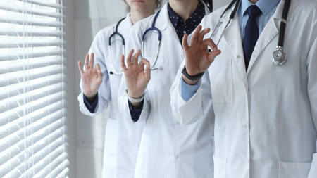 Medical team demonstrating professionalism and successful teamwork, doctors and healthcare workers in lab coats and stethoscopes showing ok sign for quality and well beingの写真素材