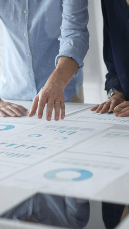 Business colleagues are examining financial data, pointing at charts and graphs spread out on a table during a collaborative analysis session in a modern office.の写真素材