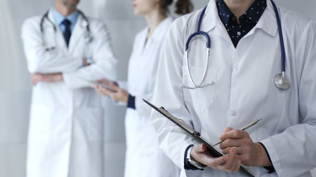 A medical professional with a stethoscope writes on a clipboard while a doctor and female assistant collaborate in a clinic, discussing patient data and healthcare informationの写真素材