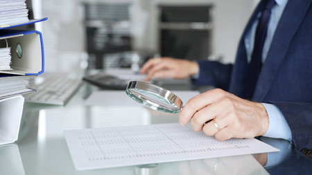 Professional male hand holding magnifying glass, looking at financial data on paper, checking details in accounting and finance. Topic of audit and taxes in businessの写真素材