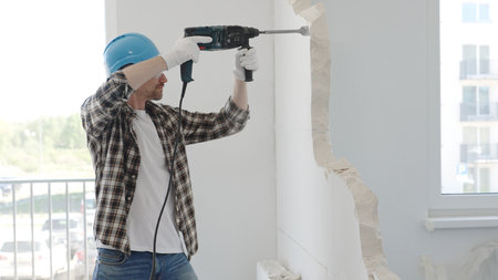 Male construction worker demolishing a wall with a hammer drill in protective gear, creating construction debris on the surrounding floor surface. Renovationの写真素材