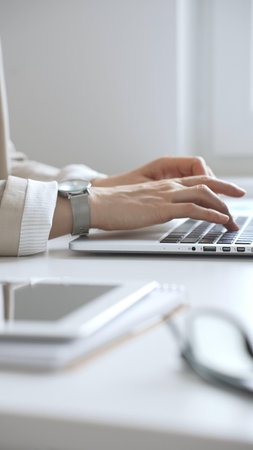 Professional businesswoman working intently on laptop, wearing tailored beige suit and silver watch,...