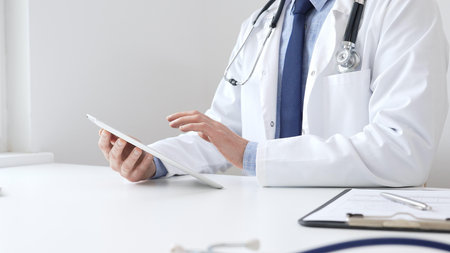 Doctor man wearing white medical coat and stethoscope using digital tablet while sitting at desk in medical office, with medical chart and pen in the foreground. Medicine conceptの写真素材