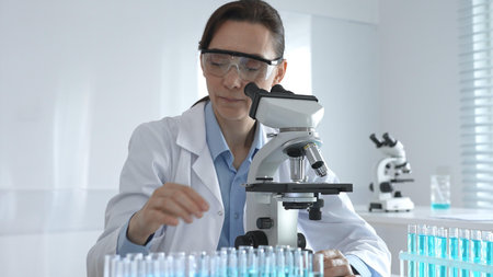 Female scientist in lab coat and safety glasses using microscope, analyzing samples in test tubes in...