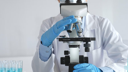 Female scientist wearing protective gear carefully adjusting microscope lens, analyzing medical research samples with precision in sterile laboratory environmentの写真素材