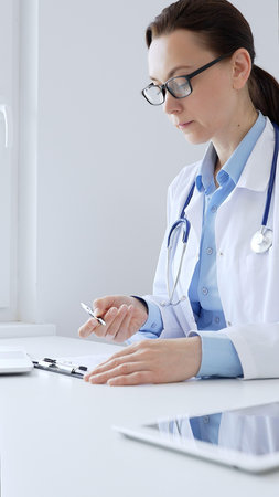 Adult female doctor completing healthcare paperwork, clipboard positioned near laptop and stethoscope in well lit clinical workspace by window. Medicine and health careの写真素材