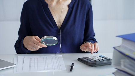 Businesswoman or auditor is analyzing financial documents while using magnifying glass and calculator at workplace desk, focused on detailed accounting review, close up. Audit and taxes in businessの写真素材