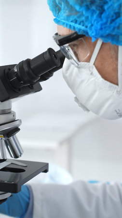 Medical scientist senior man wearing protective mask, glasses and bouffant cap using microscope doing scientific research in a laboratory. Medicine and science conceptの写真素材