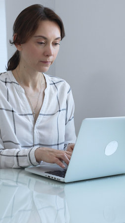 Professional businesswoman working on laptop, sitting at glass desk in bright contemporary workspace, embodying corporate success and concentration. Business people concept.の写真素材