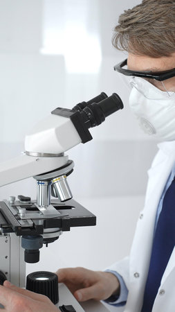 A male scientist in a protective mask looks into a microscope in a brightly lit laboratory,...
