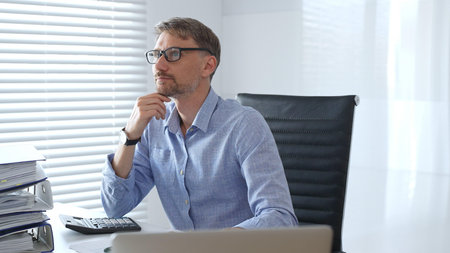 Serious male accountant is thinking while analyzing tax documents, using calculator near stacked folders, sitting in black office chair wearing crisp blue shirt. Audit and taxes in businessの写真素材