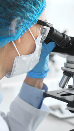 Female scientist in blue gloves and mask using microscope in laboratory setting, conducting important research and analysis, close-up. Medicine and science conceptの写真素材