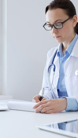 Adult doctor woman completing healthcare paperwork, clipboard positioned near laptop and stethoscope in well lit clinical workspace by window. Medicine and health careの写真素材