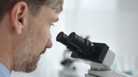 Close up of a man scientist carefully observing through a microscope in a brightly lit laboratory, conducting research for developing new medicines and treatments. Medicine and science conceptの写真素材