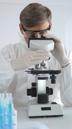 A male medical virologist in a medical gown and gloves examines viral samples under a microscope...