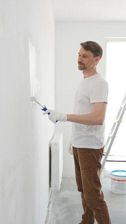Middle aged man wearing white gloves and t-shirt, is painting white wall with paint roller, experiencing home renovation and transforming living spaceの写真素材