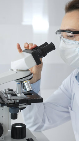 A female scientist in a protective mask and glasses carefully examines a sample under a microscope...