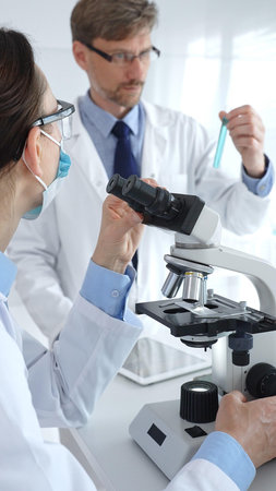 Two scientists in lab coats and safety gear conducting research, one observing through a microscope...