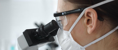 Close up of woman scientist wearing protective mask and goggles carefully examining sample through...